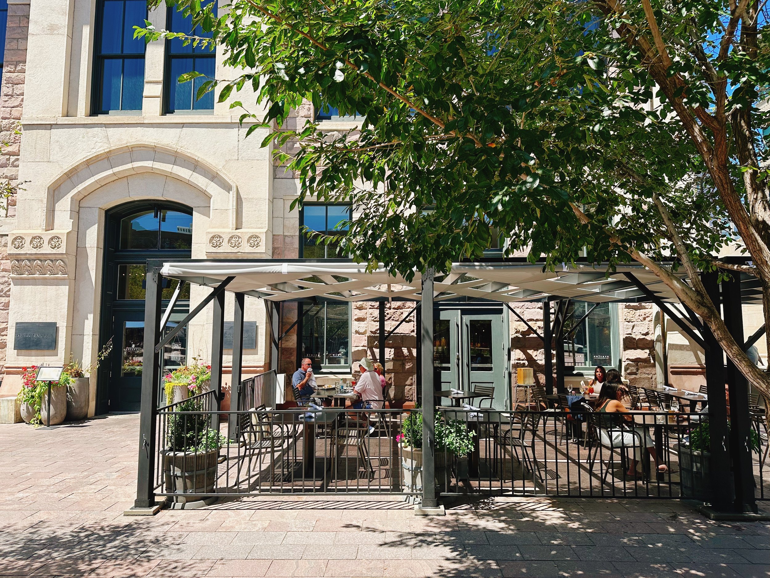 The image shows an outdoor patio area of a café or restaurant, situated in front of a historic stone building. The patio is covered with a pergola and surrounded by a black metal fence. There are several tables and chairs, with a few people sitting and enjoying their meals or drinks. Large potted plants and trees provide shade and greenery to the scene. The building has arched windows and decorative stonework.