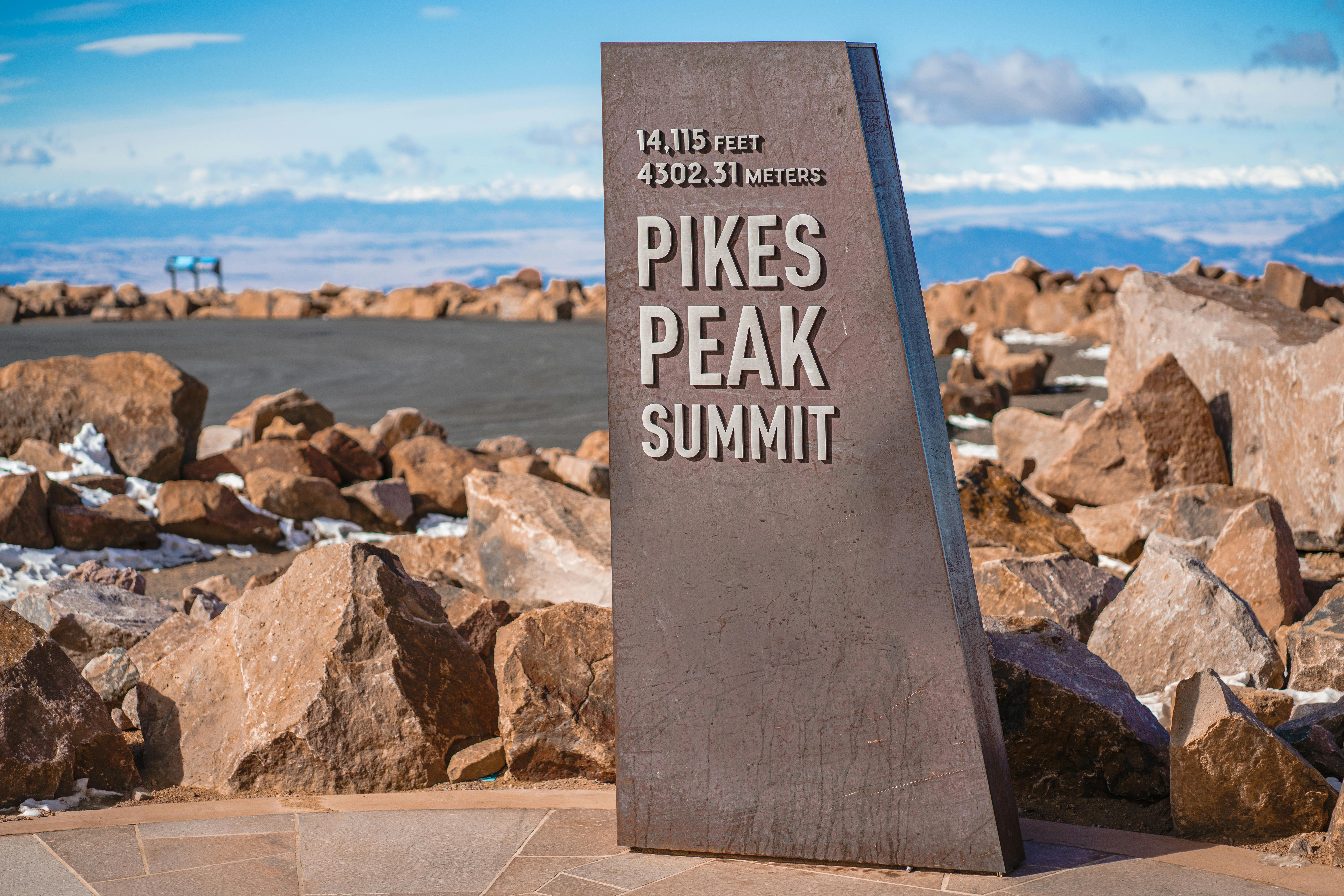 A stone marker at the summit of Pikes Peak, indicating an elevation of 14,115 feet (4,302.31 meters). The background features a rocky landscape with a clear blue sky and distant mountains.