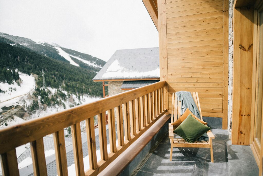 A wooden balcony with a chair that has green and orange cushions and a draped blanket. The balcony overlooks a snowy mountain landscape with pine trees and a house with a snow-covered roof.