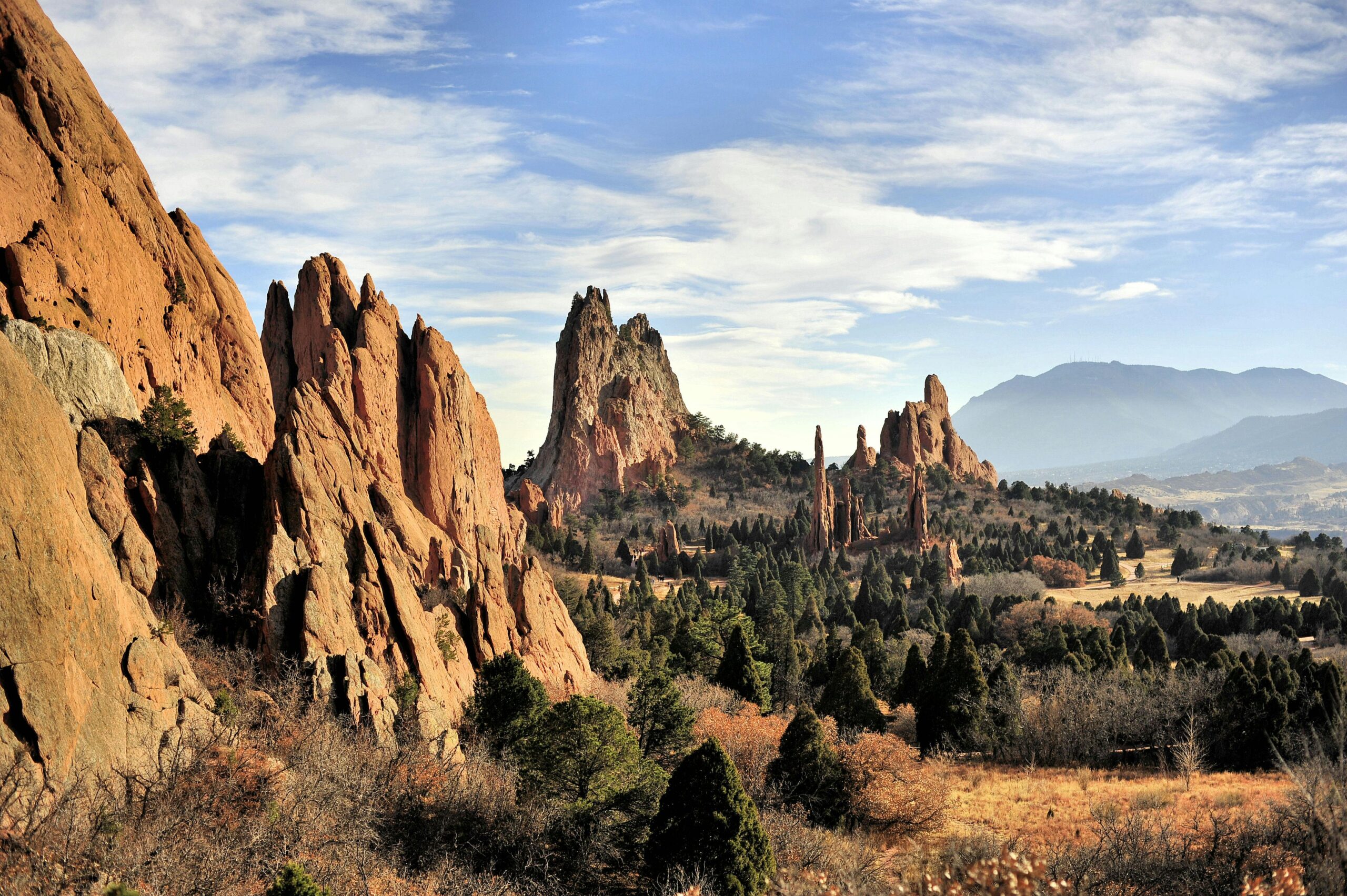 The image shows a scenic landscape featuring large, reddish rock formations with jagged peaks, surrounded by a forested area. The sky is partly cloudy, and there are distant mountains in the background. The scene is likely from a natural park or a similar outdoor setting.