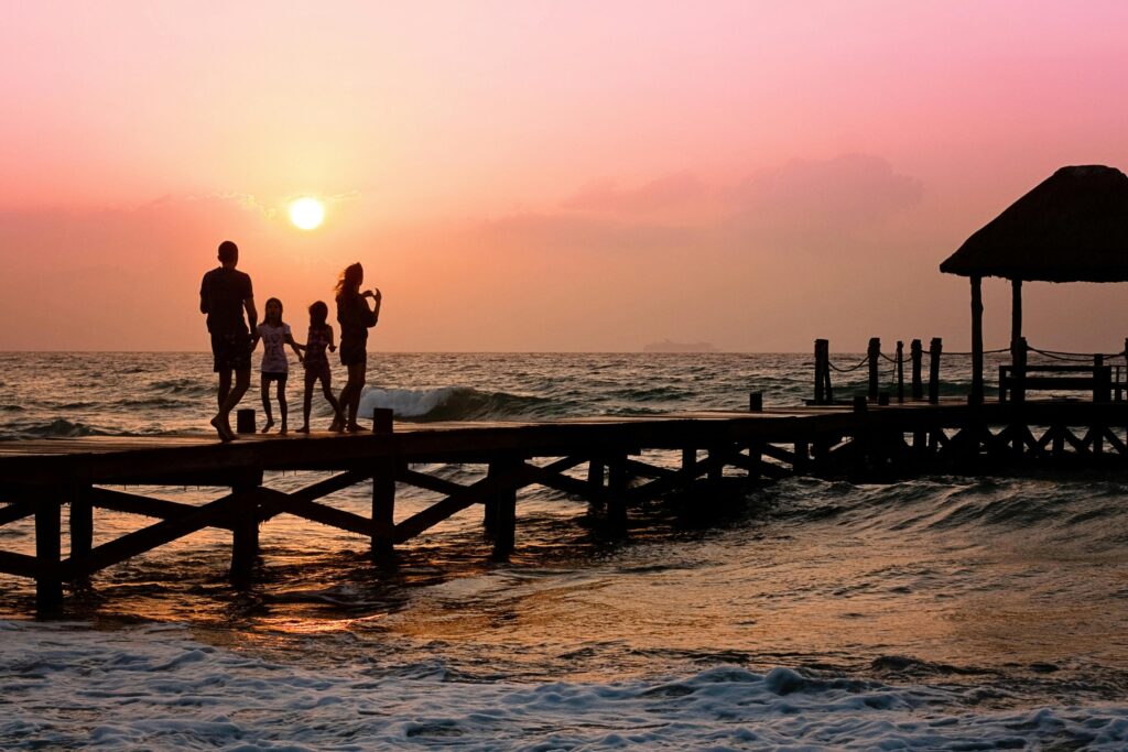 A silhouette of four people walking on a wooden pier over the ocean at sunset. The sky is a gradient of pink and orange hues, and the sun is low on the horizon. Waves are gently crashing against the pier, and there is a thatched-roof structure at the end of the pier.