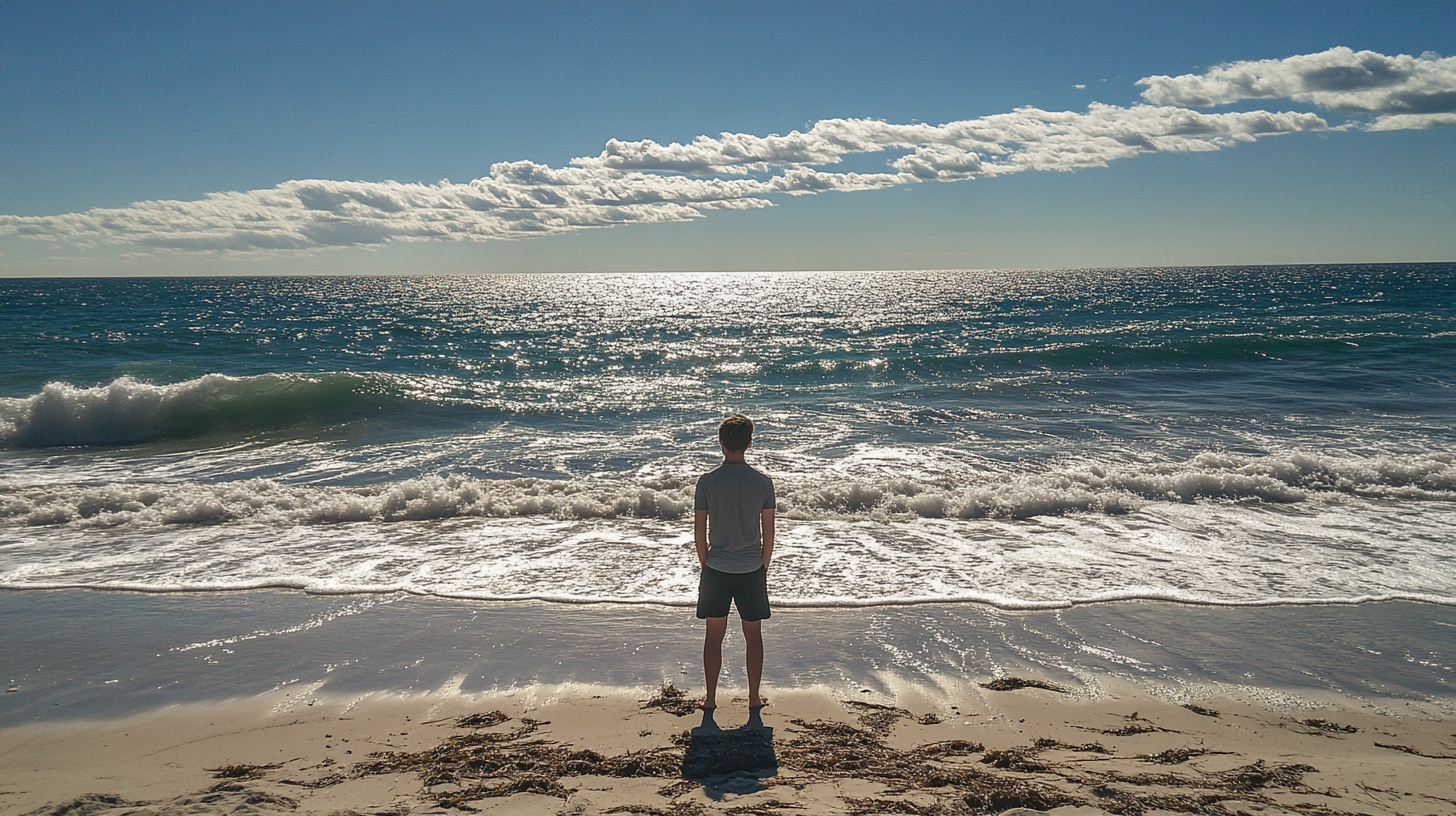 A person stands on a sandy beach, facing the ocean. The waves are gently crashing onto the shore, and the sun is reflecting off the water. The sky is clear with a few clouds.