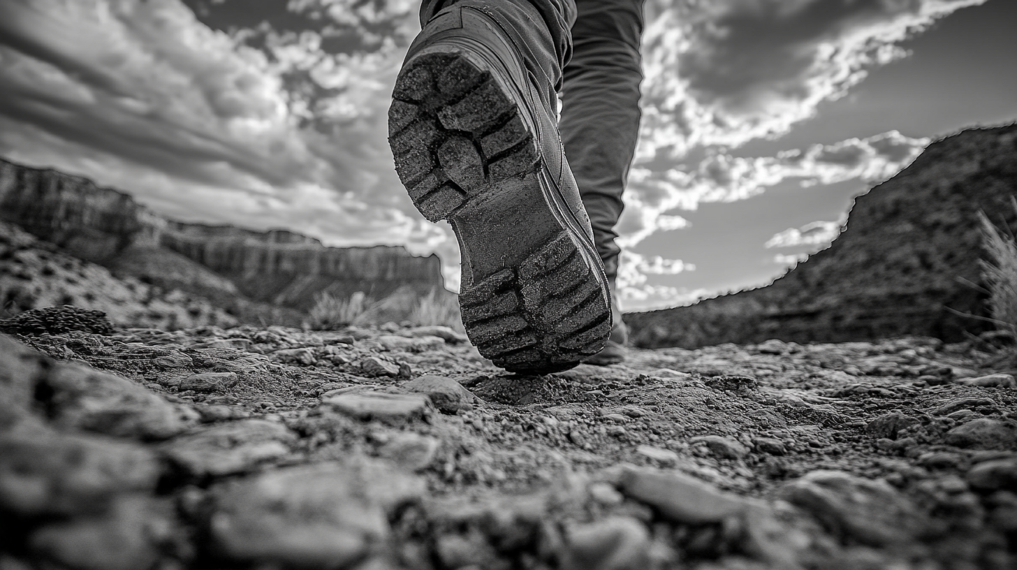 A black and white image showing a close-up of a person's boot sole as they walk on a rocky terrain. The perspective is from the ground, emphasizing the texture of the boot and the rough surface. In the background, there are blurred rock formations and a cloudy sky.